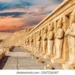 A photo of a colonnade of tall statues of standing figures in front of the Mortuary Temple of Hatshepsut at sunset. The sky is ablaze with orange, red, and purple hues, casting long shadows from the statues