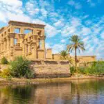 An image of the Philae Temple, a beautiful temple surrounded by water with a palm tree in the foreground. The temple is located on Agilkia Island in Egypt. Philae Temple is greco-roman and was built on an island in the Nile River.