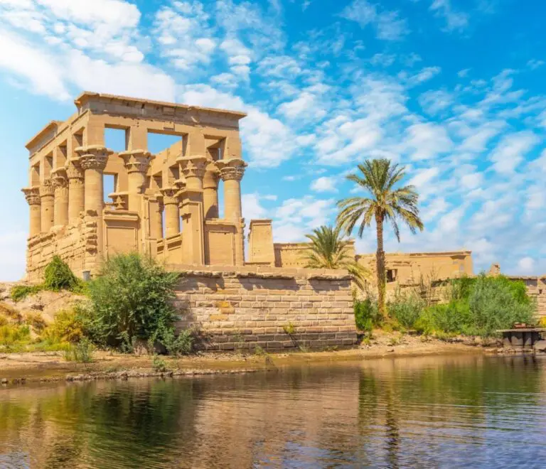 An image of the Philae Temple, a beautiful temple surrounded by water with a palm tree in the foreground. The temple is located on Agilkia Island in Egypt. Philae Temple is greco-roman and was built on an island in the Nile River.